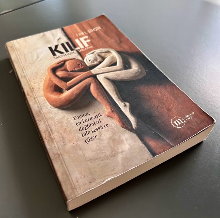A copy of the novel Kılıf lying on a table, its cover showing two intertwined figures in clay and white.