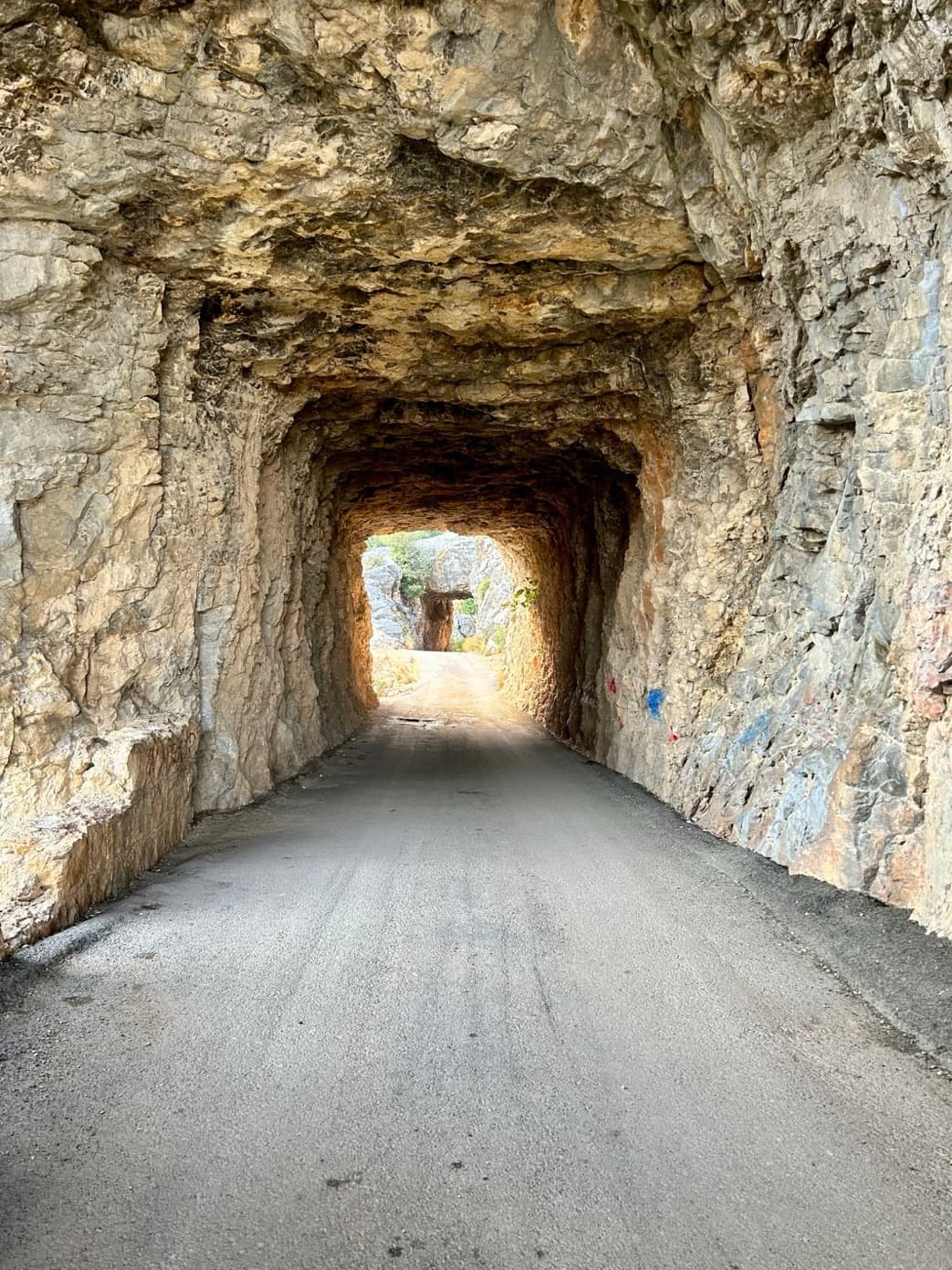 A road tunnel carved through rock, with daylight showing at the far end of the passage through the stone.